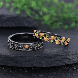 Puede incluir: Dos anillos negros con diseños de hojas y piedras preciosas. Uno presenta un patrón de hojas con piedras preciosas naranjas, el otro una banda de piedras preciosas naranjas y verdes. Los anillos están sobre un fondo oscuro.