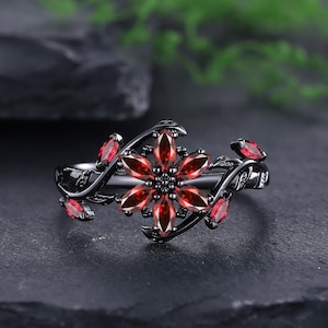 Peut inclure: Une bague en métal noir avec un motif central en forme de fleur composé de six pierres rouges taille marquise. L'anneau présente un motif de vigne, agrémenté de petites pierres rouges supplémentaires sur les côtés, créant un aspect botanique.