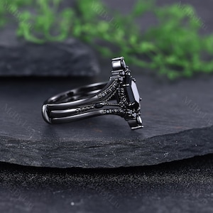 Pode incluir: Um conjunto de an&eacute;is de noivado pretos com uma pedra preta retangular e pedras pretas menores. O anel tem um design ornamentado &uacute;nico com uma banda a condizer. O conjunto &eacute; exibido numa superf&iacute;cie escura e texturizada.