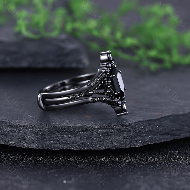 Pode incluir: Um conjunto de an&eacute;is de noivado pretos com uma pedra preta retangular e pedras pretas menores. O anel tem um design ornamentado &uacute;nico com uma banda a condizer. O conjunto &eacute; exibido numa superf&iacute;cie escura e texturizada.
