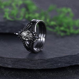 Pode incluir: Um anel preto com um design &uacute;nico. O anel apresenta uma pedra central com um padr&atilde;o semelhante a uma teia, rodeada por pedras pretas menores. A banda tamb&eacute;m &eacute; preta, criando um visual coeso e marcante. O anel est&aacute; sobre um fundo escuro.