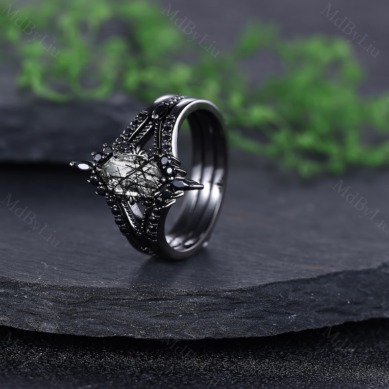 Pode incluir: Um anel preto com um design &uacute;nico. O anel apresenta uma pedra central com um padr&atilde;o semelhante a uma teia, rodeada por pedras pretas menores. A banda tamb&eacute;m &eacute; preta, criando um visual coeso e marcante. O anel est&aacute; sobre um fundo escuro.