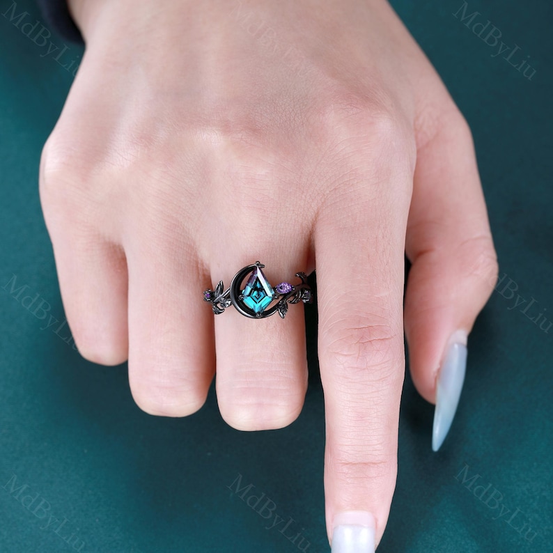 May include: Black ring featuring a teal, diamond-shaped gemstone set within a crescent moon design. The ring has small purple accent stones and floral details on the band, creating an elegant and unique piece of jewelry.