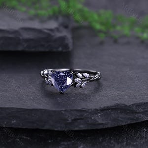 May include: A black ring with a heart-shaped, blue gemstone centre. The ring has a twisted band and is decorated with small, white gemstones either side of the heart. The ring is set against a dark background.