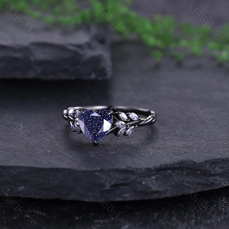 May include: A black ring with a heart-shaped, blue gemstone centre. The ring has a twisted band and is decorated with small, white gemstones either side of the heart. The ring is set against a dark background.