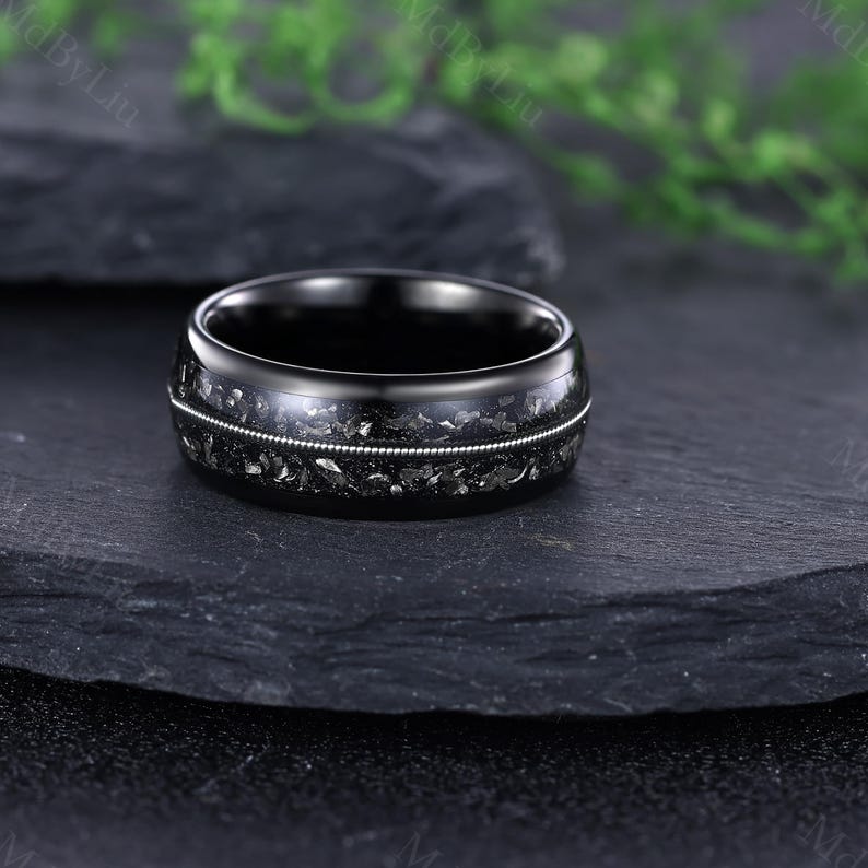 Pode incluir: Um anel preto com uma incrusta&ccedil;&atilde;o texturizada cinza escuro e uma fina faixa prateada. O anel &eacute; exibido sobre uma superf&iacute;cie de pedra cinza escuro, destacando seu design. A faixa do anel tem aproximadamente 8 mm de largura.