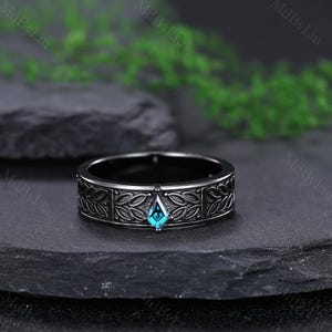 May include: A black ring with a central, teal-colored, diamond-shaped gemstone. The band features a detailed, repeating leaf pattern. The ring is displayed on a dark, textured surface, with greenery in the background.