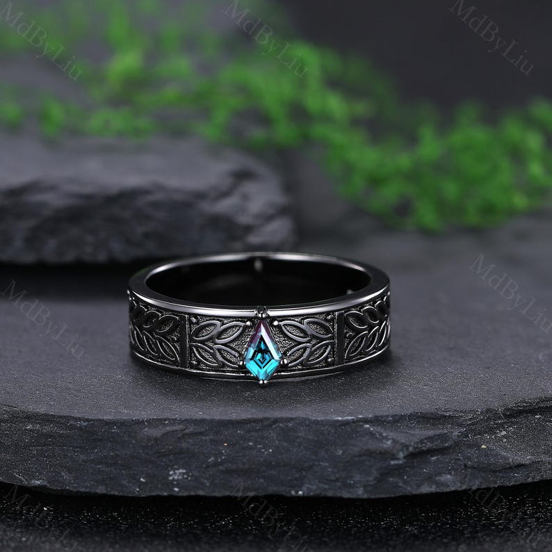 May include: A black ring with a central, teal-colored, diamond-shaped gemstone. The band features a detailed, repeating leaf pattern. The ring is displayed on a dark, textured surface, with greenery in the background.