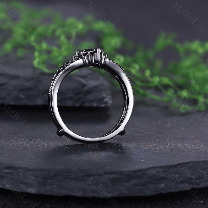 Pode incluir: Um anel de noivado preto com uma pedra preta central e pedras de destaque menores. O anel tem um design &uacute;nico com haste dividida, em um fundo escuro.