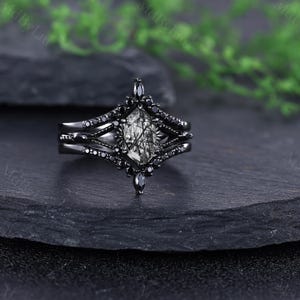 Pode incluir: Um conjunto de an&eacute;is pretos com uma pedra central de formato geom&eacute;trico com um padr&atilde;o em preto e branco. O anel &eacute; rodeado por pedras pretas menores e tem uma banda a condizer. O anel &eacute; exibido numa superf&iacute;cie escura e texturizada.
