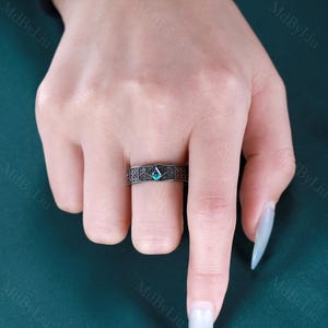 May include: A dark gray ring with an intricate floral pattern and a teal-colored, diamond-shaped gemstone. The ring is worn on a finger, showcasing the detailed design and the vibrant color of the stone.