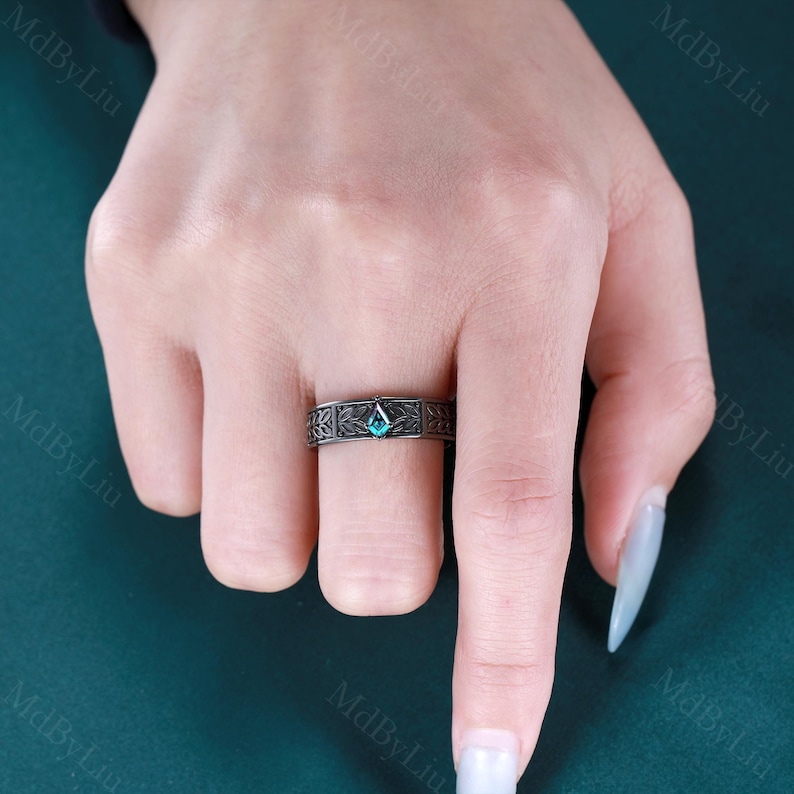 May include: A dark gray ring with an intricate floral pattern and a teal-colored, diamond-shaped gemstone. The ring is worn on a finger, showcasing the detailed design and the vibrant color of the stone.