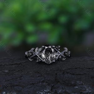 May include: Black engagement ring with a large oval-shaped gemstone and black diamond accents. The ring features a delicate leaf design on the band.