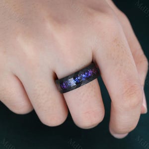 May include: A black ring with a geometric, faceted design. The ring has a colourful inlay of purple and blue, reminiscent of a galaxy. The ring is worn on a finger, highlighting its unique design and craftsmanship.
