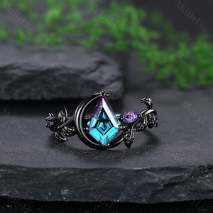 May include: A black ring with a large, teal, kite-shaped center stone. The ring features a crescent moon design and is accented with purple gemstones and leaf details. The ring is set against a dark background.