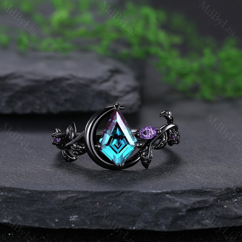 May include: A black ring with a large, teal, kite-shaped center stone. The ring features a crescent moon design and is accented with purple gemstones and leaf details. The ring is set against a dark background.