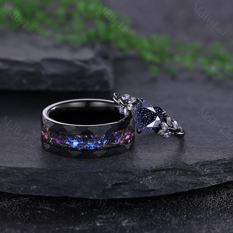 May include: Two black rings. One ring has a galaxy-like inlay with blue, purple, and pink hues. The other ring features a dark blue, heart-shaped stone and small white stones. The rings are set against a dark background.