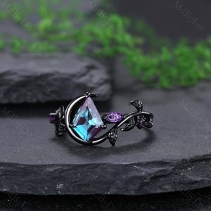May include: A black ring with a kite-shaped, teal and purple gemstone. The ring features a floral design with purple accents and a crescent moon shape around the main stone. The ring is set on a dark surface.