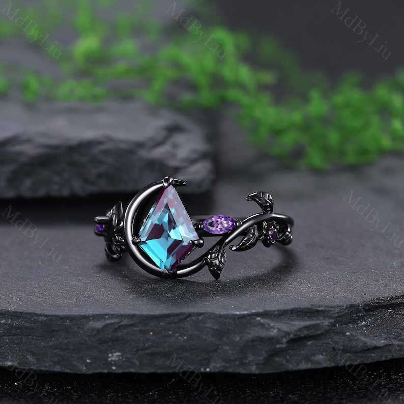 May include: A black ring with a kite-shaped, teal and purple gemstone. The ring features a floral design with purple accents and a crescent moon shape around the main stone. The ring is set on a dark surface.