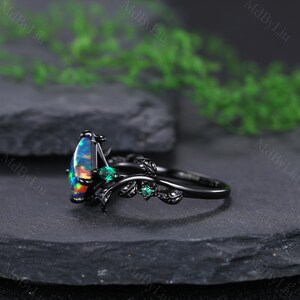 May include: Black ring featuring a pear-shaped opal with vibrant colors and small green gemstones. The ring has a nature-inspired design with black leaves and a dark band. The ring is set on a dark surface.