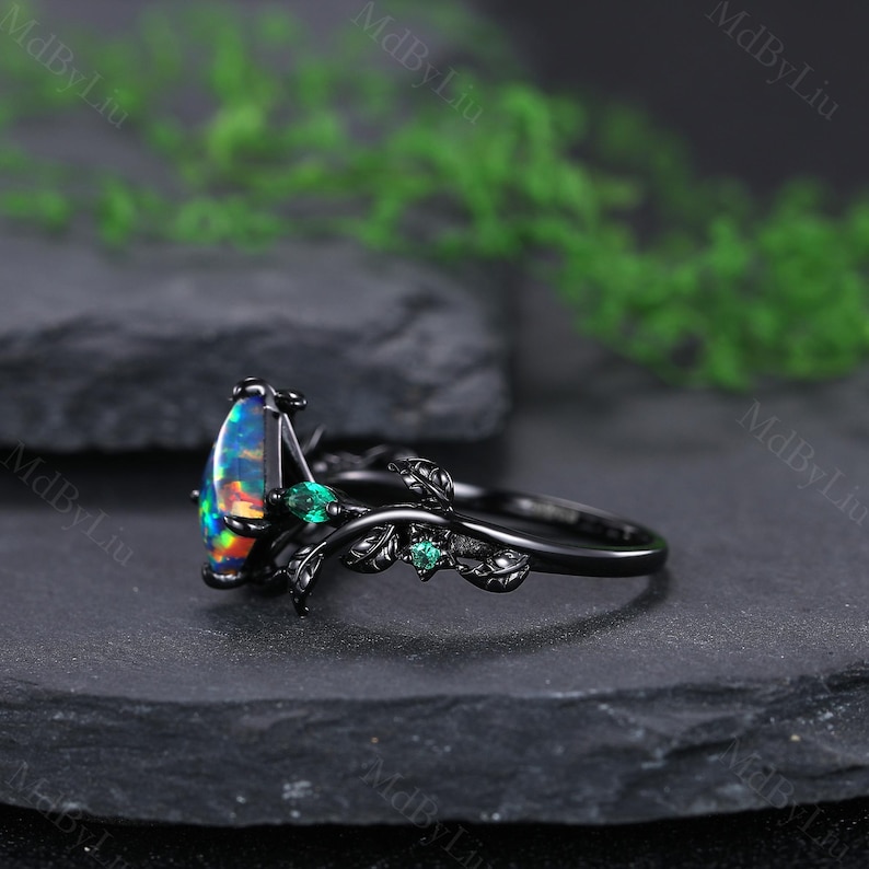 May include: Black ring featuring a pear-shaped opal with vibrant colors and small green gemstones. The ring has a nature-inspired design with black leaves and a dark band. The ring is set on a dark surface.
