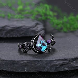 May include: Black ring featuring a large, kite-shaped, teal and purple gemstone. The ring has a crescent moon design with floral and purple accent stones. The ring is set on a dark surface.