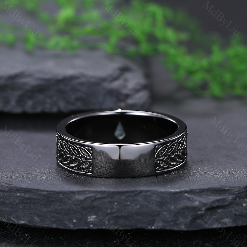 May include: A black ring with a leaf pattern engraved around the band. The ring has a polished finish and a small, light-colored stone set in the center. The ring is displayed on a dark, textured surface.