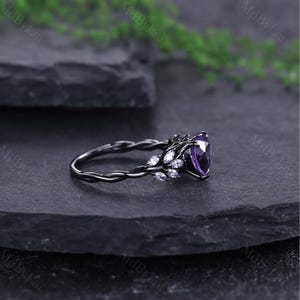May include: A black ring featuring a large, heart-shaped purple gemstone. The ring has a twisted band and is decorated with small, white accent stones. The design incorporates a floral theme, with the gemstones arranged in a leaf-like pattern.