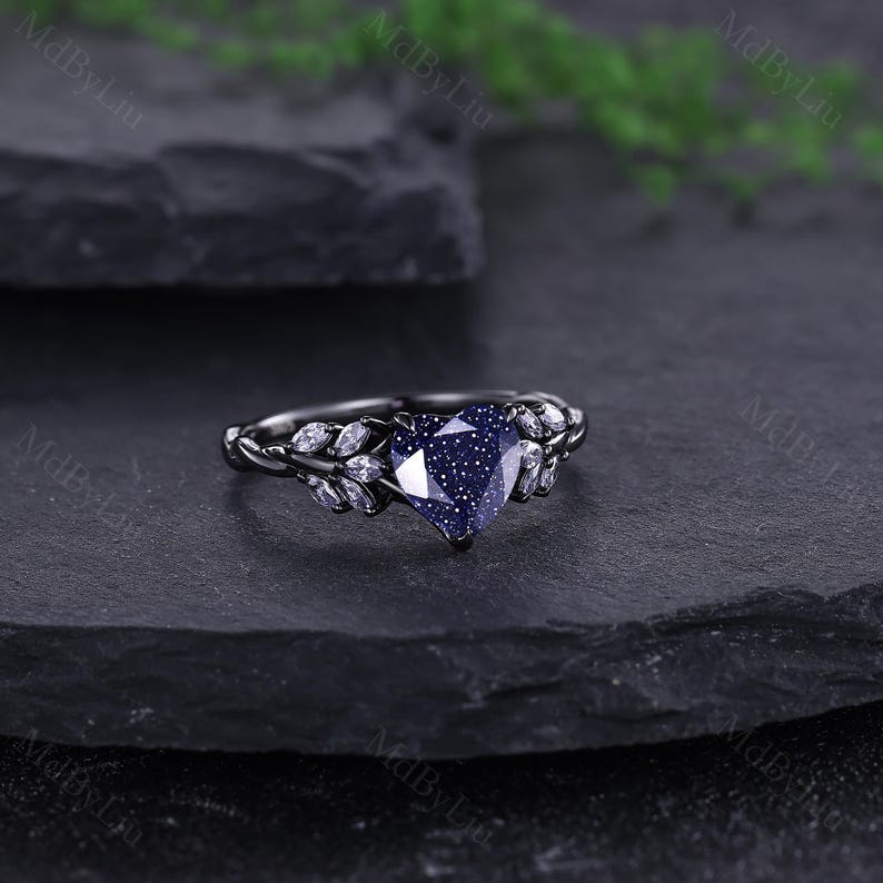 May include: A black ring featuring a heart-shaped, dark blue gemstone with a speckled appearance. The ring has a leaf design with small white gemstones. The ring is set on a dark grey surface. The ring is a piece of jewellery.