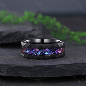 May include: A black ring featuring a geometric band design and a vibrant, galaxy-inspired inlay with shades of purple, blue, and pink. The ring is displayed on a dark surface.