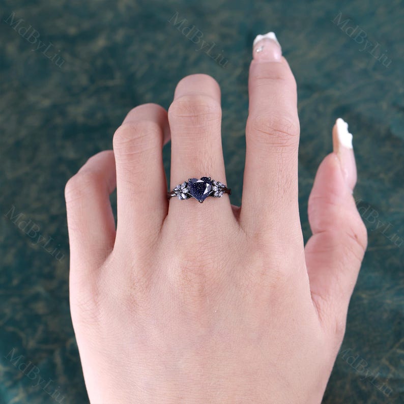 May include: A black ring with a heart-shaped, blue-speckled stone. The ring has floral detailing on the band. The ring is on a person's finger, with a blurred background.