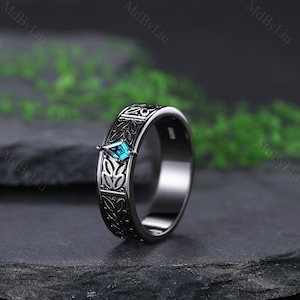 May include: A black ring with an engraved floral pattern and a teal, diamond-shaped gemstone. The ring is set against a dark background, highlighting the intricate details of the design. The gemstone adds a pop of color to the otherwise monochromatic piece.
