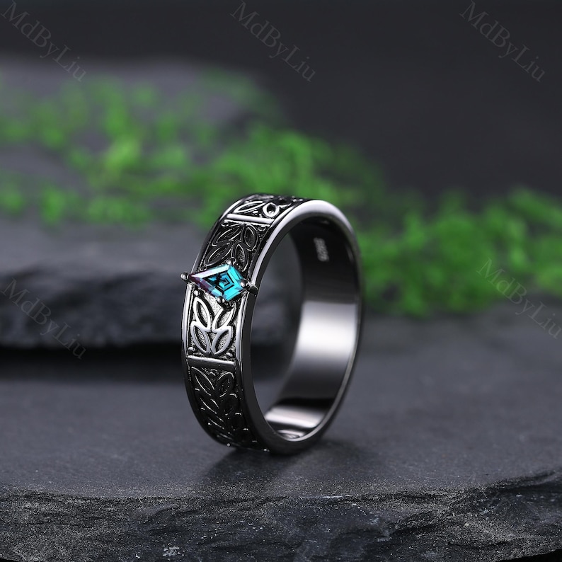 May include: A black ring with an engraved floral pattern and a teal, diamond-shaped gemstone. The ring is set against a dark background, highlighting the intricate details of the design. The gemstone adds a pop of color to the otherwise monochromatic piece.