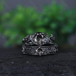 May include: A black gold engagement ring set with a black diamond and black diamond accents. The ring features a leaf design and is set on a black background.