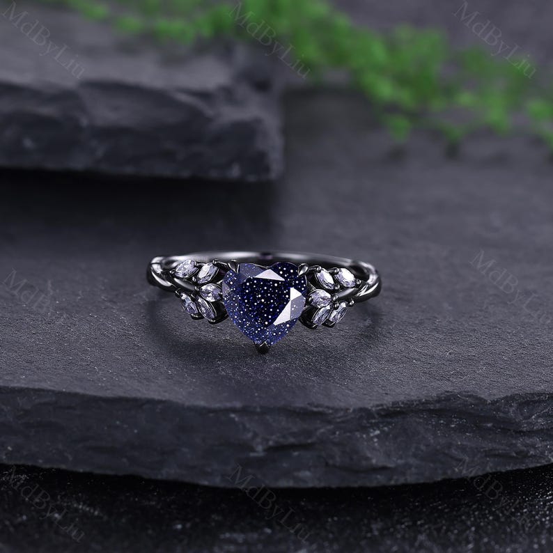 May include: A black metal ring with a heart-shaped, blue gemstone centre. The ring features leaf-like accents with small, clear stones. The ring is set against a dark, textured background.