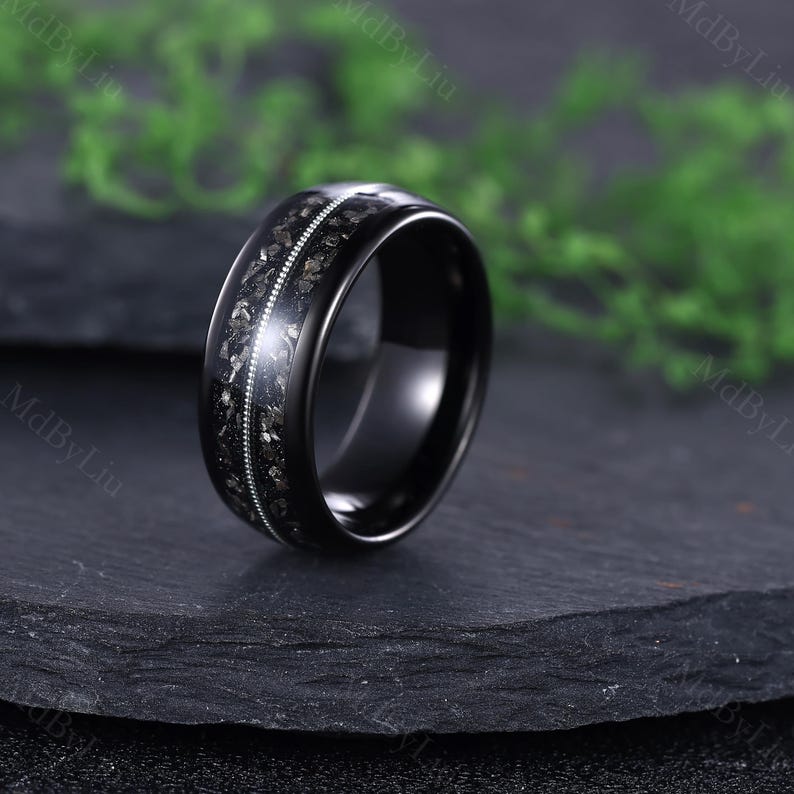 Pode incluir: Um anel preto com uma faixa central de manchas cinza escuro e prateadas, delimitada por uma fina linha de pequenas pedras brilhantes. O anel tem um acabamento polido e brilhante, sobre um fundo escuro.