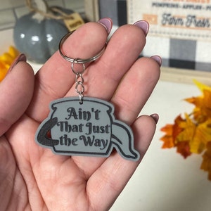 May include: Gray keychain with a cauldron shape and the text "Ain't That Just the Way" in black lettering.