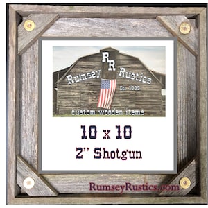 May include: A rustic wooden frame with a gray background. The frame is decorated with four bullet casings. The frame contains a printed image of a red barn with an American flag hanging from it. The image also includes the text "Rumsey Rustics Est. 1999 custom wooden items 10 x 10 2" Shotgun RumseyRustics.com".