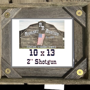 May include: A rustic wooden picture frame with a gray background and a logo for Rumsey Rustics. The logo features a red, white, and blue American flag and the text "Rumsey Rustics Est. 1999". The frame is decorated with four shotgun shell casings. The text "10 x 13 2" Shotgun" is printed on the frame.