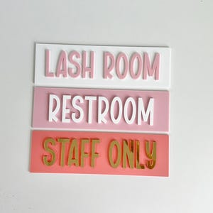 May include: Three acrylic signs with different text. The top sign is white with pink text that says "LASH ROOM". The middle sign is pink with white text that says "RESTROOM". The bottom sign is red with gold text that says "STAFF ONLY".