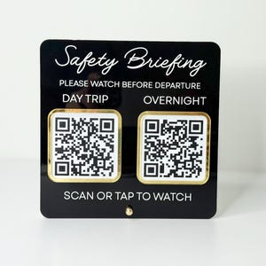 May include: Black rectangular sign with the words "Safety Briefing" in cursive script. It includes the text "Please watch before departure" and QR codes for day trips and overnight stays. The sign also says "Scan or tap to watch."