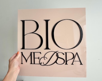 May include: A square sign with the words "BIO ME SPA" in black, elegant lettering. The sign has a light pink background. The sign is being held by a person.