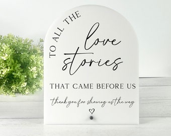 May include: White acrylic sign with a rounded top and black text that reads "To all the love stories that came before us, thank you for showing us the way." A small heart symbol is below the text.