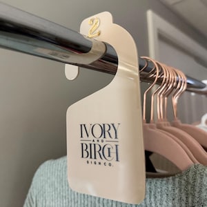 May include: A cream-colored clothing rack divider with the number "2" in gold. The divider is hanging on a chrome rod, with the "Ivory and Birch Sign Co." logo printed on it. Pink velvet hangers are visible.