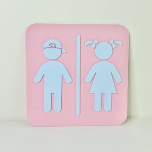 May include: A pink square sign with rounded corners features light blue silhouettes of a person wearing a baseball cap and a person with pigtails, separated by a vertical line. The sign indicates a restroom.