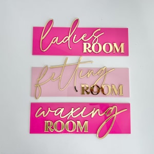 May include: Three rectangular signs with gold lettering on a white background. The top sign is hot pink with the words "Ladies Room". The middle sign is light pink with "Fitting Room". The bottom sign is hot pink with "Waxing Room".