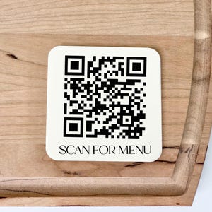 May include: A square, off-white QR code sticker with the text "SCAN FOR MENU" is placed on a wooden cutting board. The QR code is black and white, and the cutting board has a light brown color with a darker brown handle.