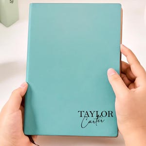 May include: A teal-colored journal with the name "TAYLOR CARTER" printed on the cover. The journal is held open by two hands. A black and gold pen rests on the white surface next to the journal. A white alarm clock and books are in the background.