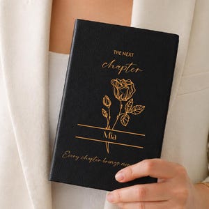 May include: A black journal with gold lettering and a rose illustration is held by a person wearing a cream-colored blazer. The journal's cover reads "THE NEXT chapter" and "Mia". The bottom of the journal reads "Every chapter brings new beginnings."