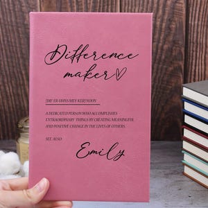 May include: A pink leather-bound journal with the words "Difference Maker" and a heart symbol. The journal also includes the definition of a difference maker and the name "Emily" at the bottom.